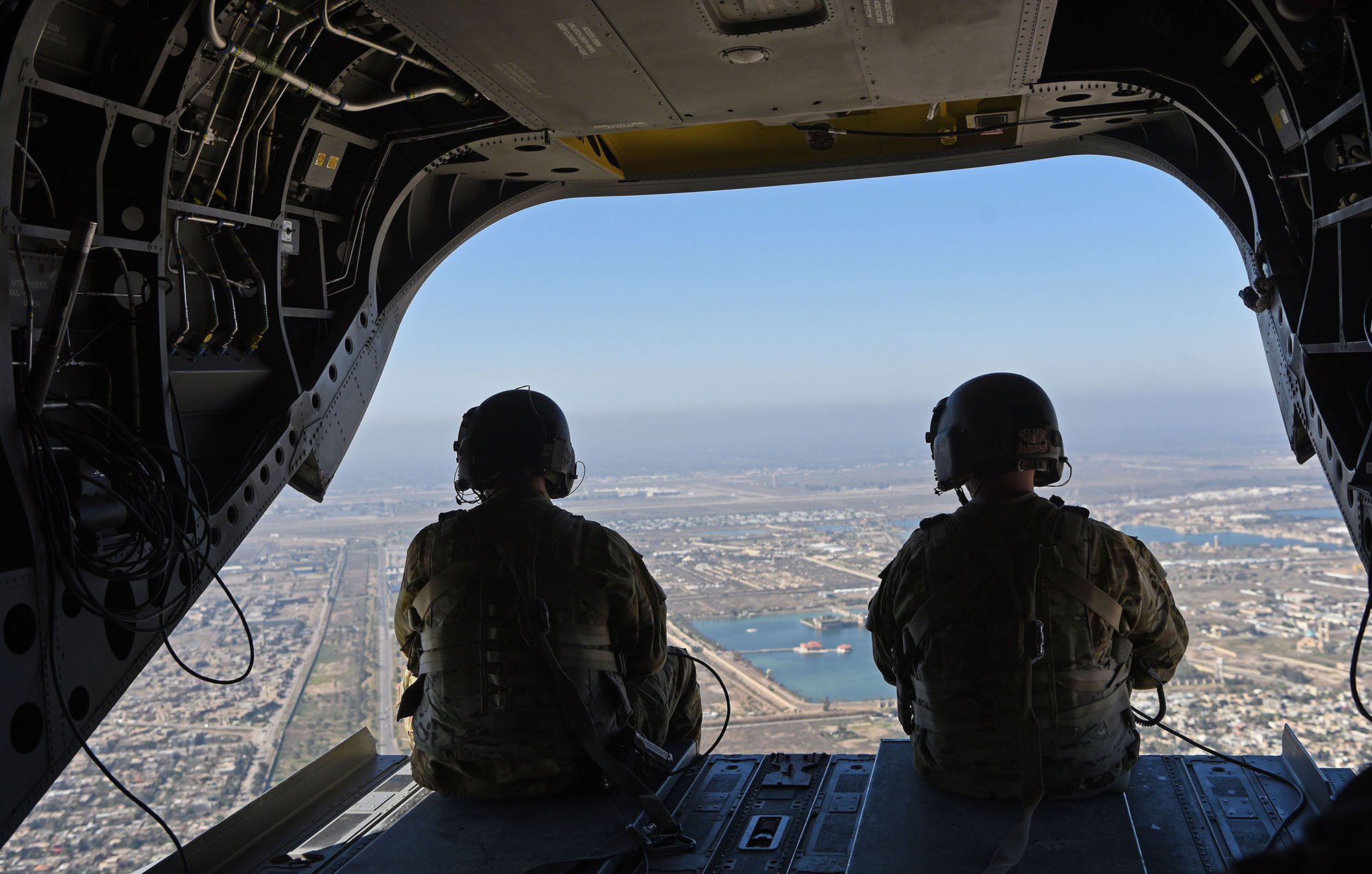 در این تصویر، دو نفر با تجهیزات نظامی در حال نشستن در یک هلیکوپتر هستند. آنها در حال مشاهده مناظر بیرون از هلیکوپتر هستند که نمایی از یک شهر و آبهای اطراف آن قابل مشاهده است. آسمان آبی و روشنی روز نشاندهنده یک روز صاف است. محیط داخلی هلیکوپتر نیز شامل تجهیزات و سیمکشیهای نظامی است.