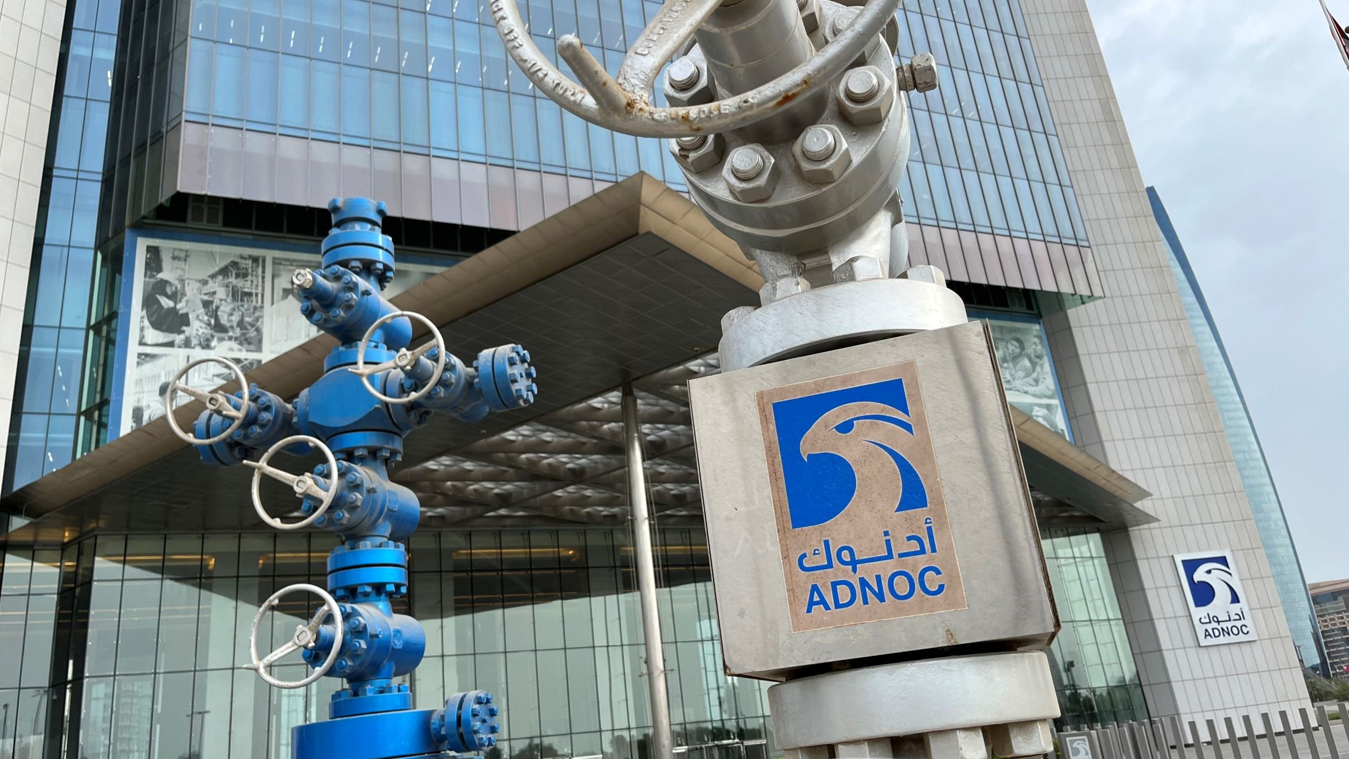 تصویر نشاندهنده نمای ساختمان شرکت نفت ابوظبی (ADNOC) است. در جلو، دو ساختار فلزی وجود دارد که شبیه به تجهیزات صنعتی و دکلهای نفتی هستند. یکی از این تجهیزات به رنگ آبی و دیگری نقرهای است. در سمت راست تصویر، لوگوی ADNOC با نوشته عربی و انگلیسی قابل مشاهده است. ساختمان دارای ظاهری مدرن و شیشهای است که عناصر طراحی معاصر را به نمایش میگذارد.