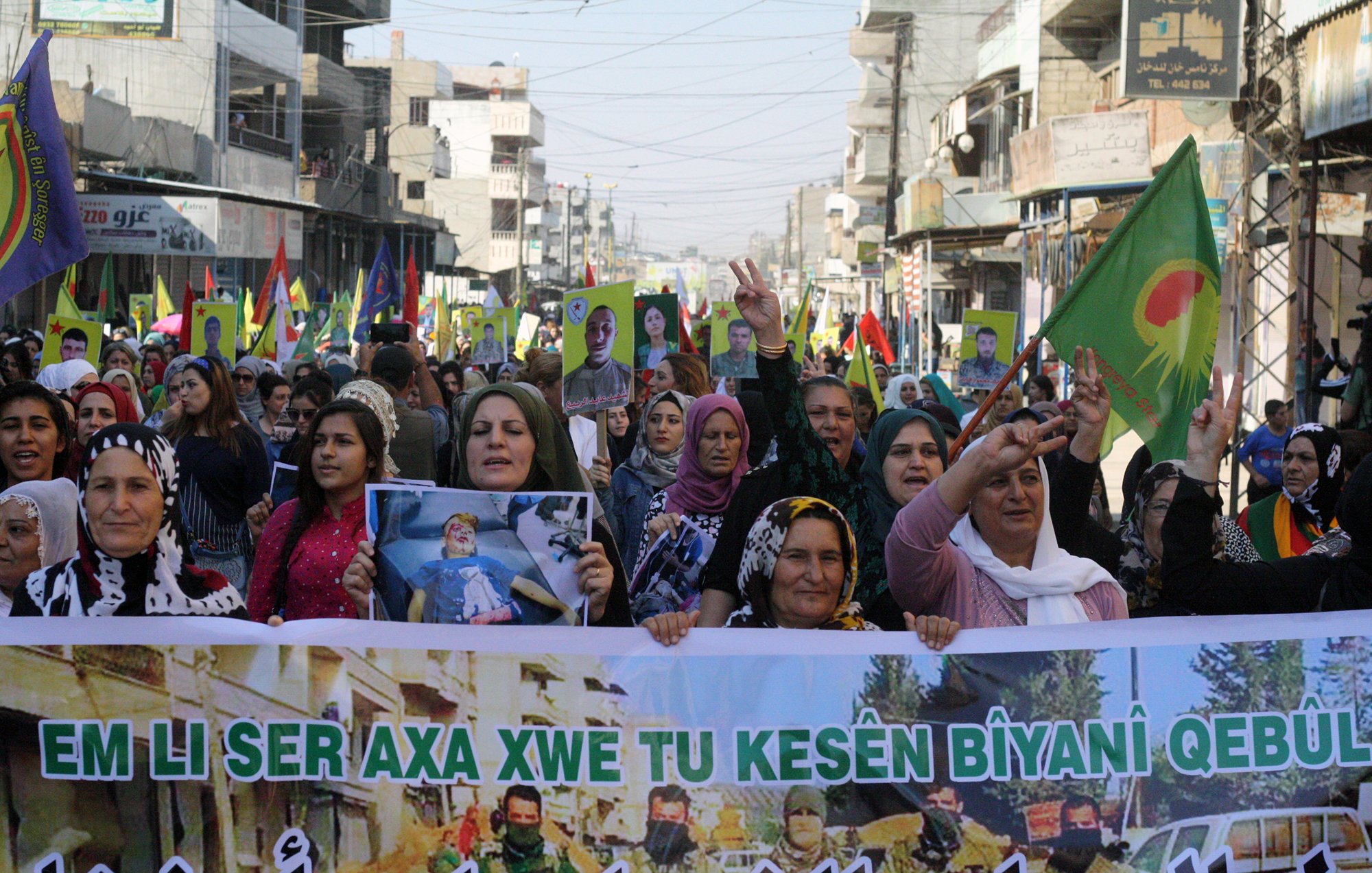 تصویر یک تظاهرات بزرگ را نشان میدهد که گروهی از زنان در آن شرکت دارند. آنها در خیابانی شلوغ راه میروند و پلاکاردهایی به دست دارند. تعدادی پرچم با نمادهایی خاص نیز در دست دارند. زنان با لبخند و دستهای بالا به نشانه حمایت و ابراز احساسات مشارکت میکنند. محیط اطراف شامل ساختمانها و جمعیت دیگری است که در پسزمینه دیده میشود. این تصویر به وضوح نشاندهنده یک حرکت اجتماعی یا سیاسی است.