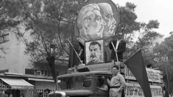 تصویر نشاندهنده یک خودروی قدیمی است که در یک مراسم راهپیمایی یا تظاهرات شرکت کرده است. بر روی ماشین بزرگنمایی از چهرههای افرادی با موهای کوتاه و مقدار زیادی نماد و پرچمهایی وجود دارد. فردی در حالت ایستاده بر روی خودرو قرار دارد و به نظر میرسد که در حال صحبت یا اشاره به تماشاگران است. پسزمینه شامل گروهی از مردم است که در کنار خیابان ایستادهاند، و فضایی از شور و اشتیاق را در این رویداد به تصویر میکشد.