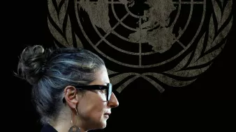 زنی با موهای خاکستری و عینک، در کنار نماد سازمان ملل، در حال صحبت کردن است.