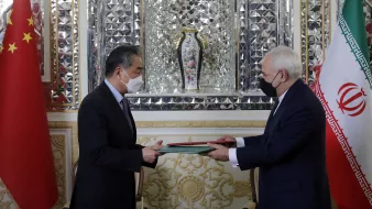 در این تصویر، دو دیپلمات در حال تبادل یک سند مهم هستند. در سمت چپ پرچم چین و در سمت راست پرچم ایران قرار دارد. هر دو فرد ماسک بر صورت دارند و بهخاطر شیوع بیماریهای تنفسی احتیاط میکنند. میز که بر روی آن سند قرار دارد، بهنظر زیبا و مزین است. پسزمینه شامل تزئینات پیچیده و یک گلدان نیز هست که به فضای رسمی و دیپلماتیک این دیدار افزوده است.