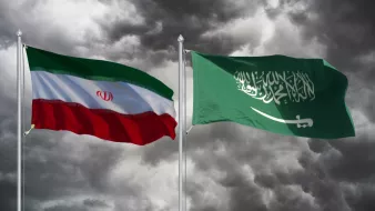 در تصویر، دو پرچم به نمایش درآمدهاند. پرچم چپ، پرچم ایران است که شامل رنگهای سبز، سفید و قرمز به صورت افقی و نشان هشتپر در وسط آن است. پرچم راست، پرچم عربستان سعودی است که رنگ اصلی آن سبز تیره است و در وسط آن نوشته عربی به رنگ سفید وجود دارد. زمینه تصویر ابری و طوفانی به نظر میرسد که حسی از تنش و درگیری را منتقل میکند.