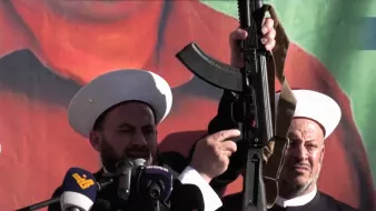 در این تصویر، دو فرد که در حال سخنرانی هستند، به نظر میرسد در یک مراسم یا تجمع حضور دارند. یکی از آنها سلاحی را در دست دارد و دیگر فرد در سمت راست نیز در حال نظارت یا همراهی با او است. پسزمینه تصویر شامل رنگهای زنده و گرافیکی است. هر دو فرد کلاههای مخصوص خود را بر سر دارند که نشاندهنده هویت مذهبی یا فرهنگی آنها است. احساس کلی تصویر به نظر میرسد جدی و تأکیدی بر موضوعی مهم باشد.