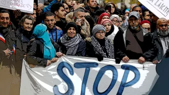 تصویر یک تجمع اعتراضی را نشان میدهد که در آن افراد مختلفی در حال ایستادن هستند. آنها یک بنر بزرگ را در دست دارند که روی آن کلمه "STOP" نوشته شده است. جمعیت شامل مردان و زنان با پوششهای متفاوت است، و به نظر میرسد که آنها خواسته یا نگرانی خاصی را بیان میکنند. پسزمینه شلوغ و چندنفره است که احساس اتحاد و جنبش اجتماعی را ایجاد میکند.