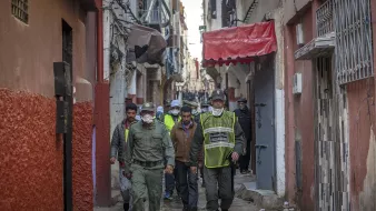 در این تصویر، یک کوچه باریک و قدیمی دیده میشود. در دو طرف کوچه، ساختمانها با رنگهای مختلف نمایان هستند. گروهی از افرادی که لباسهای فرم دارند، در حال قدم زدن در کوچه هستند. برخی از آنها کلاه و ماسک به چهره دارند. محیط به نظر شلوغ و فعال میرسد و افراد مشغول به انجام کاری هستند. این تصویر فضایی محلی و اجتماعی را به نمایش میگذارد.