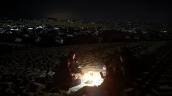 در این تصویر، یک گروه از افراد در شب در حال دور هم نشستن در بیابان هستند. آنها در اطراف آتش نشستهاند و نور ملایم آتش نور را بر چهرههایشان میتاباند. در پسزمینه، میتوان چادرها و نورهای دیگری را که در دوردست قرار دارند، مشاهده کرد. این صحنه حس صمیمیت و نزدیکی به طبیعت را منتقل میکند. همچنین، فعالیتهای شبانه و سکوت بیابان به فضای تصویر عمق بیشتری میبخشد.