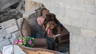 مادری بر روی مبل خراب نشسته و موهای کودک را نوازش میکند. در پسزمینه، ویرانی مشهود است.