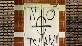 در این تصویر، درب یک ساختمان با رنگ سیاه نوشته شده است. در وسط درب علامت دایرهای با خطی عمودی و افقی وجود دارد که به همراه نوشته "NO ISLAM!" که به معنای "اسلام نه!" است، روی درب دیده میشود. این نوشته به روشنی یک پیام اعتراضی یا نفرتآمیز را منتقل میکند. زمینه دیوار نیز آجری و با رنگهای متفاوت است.