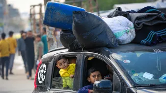 در این تصویر، یک خودرو دیده میشود که باری بر روی سقف آن قرار دارد. تعدادی کیسه و لباس به شکل نامنظم روی سقف خودرو گذاشته شدهاند. درون خودرو، دو کودک نشستهاند؛ یکی از آنها در حال نگاه کردن به بیرون از پنجره است و دیگری به سمت او توجه دارد. در پسزمینه، افرادی در حال حرکت هستند و فضایی شلوغ و شهری به نظر میرسد. وضعیت کلی تصویر نشاندهنده زندگی روزمره در یک محیط حاشیهای است.