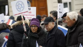 در این تصویر، گروهی از افراد در حال اعتراض هستند. آنها پلاکاردها و تابلوهای مختلفی در دست دارند. برخی از افراد لباسهای گرم به تن دارند و در میان آنها یک زن با عینک دیده میشود. تابلوها شامل پیامهایی مرتبط با اسرائیل هستند و برخی از افراد به سمت دوربین نگاه میکنند. فضای تجمع در یک مکان عمومی به نظر میرسد.