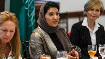 در این تصویر، یک جلسه یا گردهمایی در حال برگزاری است. در مرکز تصویر، زنی با حجاب و لباسی رسمی نشسته است و به صحبت کردن مشغول است. در سمت چپ، زنی دیگر با موی بلند و لباسی غیررسمی دیده میشود که به نظر میرسد به سمت صحبت کننده در حال گوش کردن است. در سمت راست، زنی دیگر با لباسی رسمی و لباس خط دار نشسته است. برخی از میوهها و نوشیدنیها روی میز قرار دارند و به نظر میرسد مهمانی یا جلسهای دوستانه برگزار میشود. پسزمینهی تصویر نیز نشاندهندهی فضایی رسمی است.