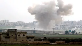 تصویر یک صحنه جنگی را نشان میدهد که در آن دود و گرد و غبار ناشی از یک انفجار دیده میشود. در پسزمینه، ساختمانهای شهری و یک منطقه باز مشاهده میشود. آسمان ابری است و حالت کلی تصویر به موقعیتی پر تنش و خطرناک اشاره دارد. دود در حال بلند شدن از زمین به سمت آسمان است و نشانههایی از ویرانی و نبرد در تصویر مشهود است.