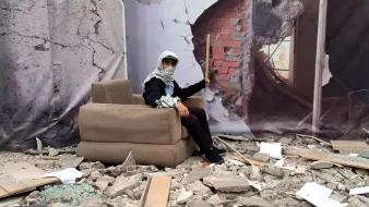 تصویر یک نوجوان یا جوان را نشان میدهد که در یک محیط تخریبشده نشسته است. او بر روی یک مبل خاکی و پوشیده از گرد و غبار نشسته و به نظر میرسد که در یک مکان ویران قرار دارد که دیوارهای آن ترک خورده و بخشی از آنها خراب شده است. در پسزمینه، تصویری از دیواری که در حال فرو ریختن است، به چشم میخورد. جوان همچنین یک یا تعدادی چوب در دست دارد و لباسهایش شامل روسری است. فضای کلی تصویر نشاندهنده ناامیدی و ویرانی است.