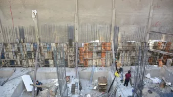 تصویر یک محل construction (ساخت و ساز) را نشان میدهد. در این تصویر، یک سازه بتونی در حال انجام است که دیوارهای آن با میلههای فولادی و قالبهای چوبی محصور شدهاند. عدهای از کارگران در حال فعالیت و جابجایی مصالح هستند. زمین پروژه نیز برخی مصالح و تجهیزات ساختمانی مانند تخته، میلههای فولادی و ابزارهای دیگر را نشان میدهد. دیوار پشتی با یک سطح صاف و عاری از هرگونه تزئینات، نمایان است.