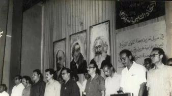 این تصویر مربوط به یک تجمع یا کنفرانس سیاسی به نظر میرسد. در پسزمینه، تابلوهایی از چهرههای معروف تاریخی یا سیاسی وجود دارد. افرادی که در جلوی صحنه ایستادهاند، موضع جدی و متفکرانهای دارند و در حال گوش دادن به سخنرانی یا برنامهای هستند. فضای کلی تصویر به نظر میرسد که تمرکز بر مسائل اجتماعی و سیاسی داشته باشد. دیوار پشتشان نیز با متنی به زبان عربی تزئین شده است.