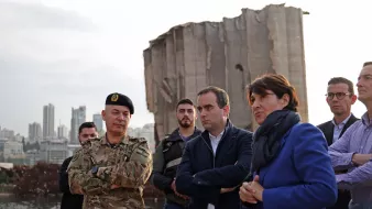 در این تصویر، گروهی از افراد در کنار یک ساختمان ویران، که ظاهراً بخشی از آن تخریب شده، ایستادهاند. یک نظامی با لباس نظامی و کلاه فرماندهی در سمت چپ قرار دارد. کنار او مردان و زنانی با لباسهای رسمی و غیررسمی ایستادهاند و به نظر میرسد که در حال صحبت و تبادل نظر هستند. پسزمینه تصویر نشاندهنده شهری با ساختمانهای بلند است. نور در تصویر کم است و فضای کلی حالت جدی و تاملی دارد.