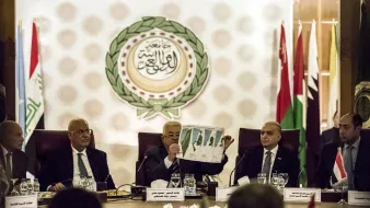 در این تصویر، گروهی از افراد در یک جلسه رسمی نشستهاند. یکی از افراد در حال نشان دادن یک نقشه یا نمودار به دیگران است. پشت آنها پرچمهای کشورهای مختلف و نماد یا لوگوی اتحادیه عرب قابل مشاهده است. فضای جلسه حرفهای و سازمانیافته به نظر میرسد. اعضای حاضر به دقت به موضوع مورد بحث گوش میدهند.