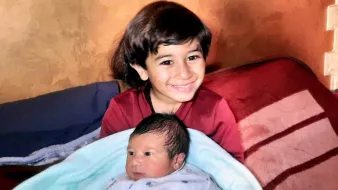 تصویر یک پسر بچهی حدود ۵ ساله را نشان میدهد که لبخند میزند و در آغوش او یک نوزاد خوابیده است. پسر بزرگتر لباس قرمز پوشیده و نوزاد در یک پتوی آبی پیچیده شده است. پسزمینه تصویر رنگی گرم دارد.