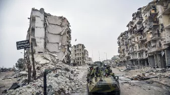 تصویر نشاندهندهٔ یک منطقهٔ ویران شده است که در آن چند ساختمان تخریب شده و در حال فرو ریختن به نظر میرسند. در این صحنه، یک وسیله نقلیه نظامی در حال حرکت است و تعدادی سرباز در آن نشستهاند. بقیهٔ محیط شامل زبالهها و نشانههای ویرانی است که به وضعیت بحرانی منطقه اشاره دارد. هوا ابری و تیره به نظر میرسد و فضایی خالی و ساکت را ایجاد کرده است.