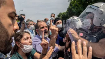 تصویر نشاندهنده یک صحنه اعتراضی است که در آن عدهای از معترضان با لباسهای غیررسمی و ماسک در حال گفتگو و مقابله با نیروهای پلیس هستند. یکی از افراد در وسط به شدت و با دستانی انداخته شده به سمت نیروهای پلیس اشاره میکند. نیروهای پلیس در حالت آمادهباش و با تجهیزات حفاظتی در برابر معترضان قرار دارند. فضایی پرتنش و اضطرابآور حاکم بر صحنه است. در پسزمینه، دیگر معترضان و تماشاچیان نیز دیده میشوند.