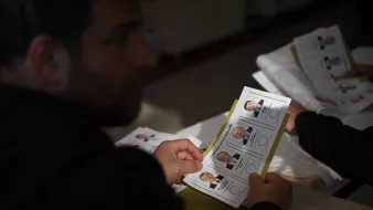 در تصویر، فردی دیده میشود که در حال بررسی یک برگه است. این برگه شامل تصاویری از چند نفر با نامها و اطلاعات مرتبط با آنهاست. احتمالاً این برگه به انتخابات یا رأیگیری مربوط میشود. فرد در حال تأمل است و در محیطی شبیه به یک مرکز رأیگیری قرار دارد. نور به آرامی به صحنه میتابد و بر تمام جزئیات تأثیر میگذارد.
