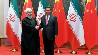 در این تصویر، دو فرد در یک فضای رسمی دیده میشوند که در حال دست دادن هستند. یکی از آنها در لباس سنتی ایرانی (شاید یک روحانی) و دیگری در لباس رسمی دیده میشود. در پسزمینه، پرچمهای ایران و چین به صورت متناوب قرار دارند که نشانهگزاری از روابط دو کشور است. فضای کلی تصویر نشاندهنده یک مراسم دیپلماتیک و همکاریهای بینالمللی است.