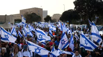 در تصویر، جمعیتی بزرگ در حال تجمع هستند و در دست دارند پرچمهای اسرائیل. بسیاری از افراد در حال صحبت و عکسبرداری با تلفنهای همراه خود هستند. فضای عمومی شلوغ و پرجنبوجوش به نظر میرسد و در پسزمینه نیز ساختمانهای دولتی یا تاریخی قابل مشاهده است. این تجمع احتمالاً مربوط به یک رویداد اجتماعی یا سیاسی است.