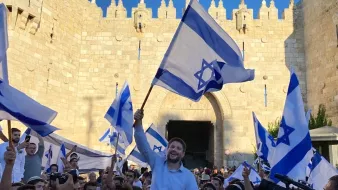 تصویر نشاندهنده جمعیتی است که با پرچمهای اسرائیل در حال جشن و شادی هستند. در میان این جمعیت، فردی در حال بلند کردن پرچم و ابراز شادی است. پسزمینه تصویر یک دروازه تاریخی و دیوار سنگی نمایان است که نشاندهنده یک مکان مهم است. افراد حاضر در تصویر لباسهای مختلفی به تن دارند و فضای کلی عکس نشاندهنده شور و نشاط است.