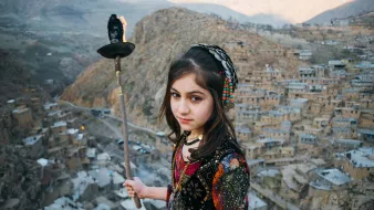 تصویر یک دختر جوان را نشان میدهد که در یک محیط کوهستانی و تاریخی ایستاده است. او لباس سنتی رنگارنگی به تن دارد و در دستش مشعل یا شعلهای در حال سوختن را نگهداشته است. در پسزمینه، یک روستای تاریخی با خانههای سنگی و دیوارهای کاهگلی در دامنه کوه نمایان است. فضای تصویر احساس اصالت و سنتهای فرهنگی را منتقل میکند. دختر با نگاه محکم و جدی به دوربین نگاه میکند، که توجه بیننده را جلب میکند.
