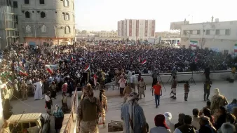 در این تصویر، یک تجمع یا راهپیمایی بزرگ در فضای باز مشاهده میشود. مردم زیادی در مقابل یک ساختمان قرار دارند و برخی از آنها پرچمهایی به دست دارند. در پسزمینه، ساختمانی بزرگ و نیمهساز دیده میشود، و ساختمانهای دیگری نیز در دوردستها وجود دارد. نور خورشید به صورت عمودی به این منطقه میتابد و جو عمومی نشاندهنده شور و هیجان مردم است. گروهی از افراد در جلو و عدهای دیگر در پشت آنها نشستهاند یا ایستادهاند.