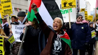 در این تصویر، گروهی از معترضان در یک راهپیمایی را مشاهده میکنیم. یک زن با پوست تیره در پیشزمینه قرار دارد که با فریاد و دست بالای خود، در حال بیان اعتراضاتش است. او پرچمهای فلسطینی و نمادهای حمایت از حقوق فلسطینیها را در دست دارد. دیگر افراد نیز در پسزمینه با پلاکاردهایی در حمایت از فلسطین و علیه صهیونیسم ایستادهاند. فضای عمومی احساس قوی از همبستگی و اعتراض را منتقل میکند.