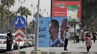 در این تصویر، خیابانی مطرح شده و در آن دو نفر در کنار پیادهرو ایستادهاند. در پسزمینه، چندین خودرو پارک شده و همچنین یک بیلبورد بزرگ با تصاویر و متنی به زبان عربی دیده میشود. این بیلبورد به احتمال زیاد به مقامات و موضوعات سیاسی اشاره دارد. تبلیغاتی برای یک برند معروف تنقلات نیز در بالای تصویر دیده میشود. در مجموع، تصویر حال و هوای شهری را به نمایش میگذارد.