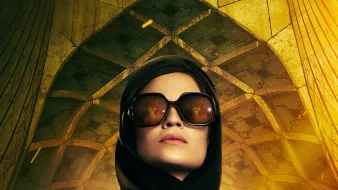 این تصویر یک زن را نشان میدهد که با پوشش سیاه و عینک آفتابی بزرگ ایستاده است. او در پسزمینهای با معماری منحصربهفرد و الگوهای هندسی قرار دارد که حال و هوای مرموز و جذابی ایجاد میکند. زن در دستانش یک سلاح کمری دارد و این به او ظاهری قوی و مصمم میدهد. نورهای زرد و نارنجی در تصویر احساس هیجان و تنش را تقویت میکنند.