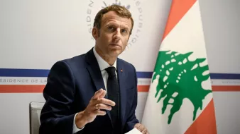 در تصویر، یک مرد با موی تیره و لباس رسمی (کت و شلوار) دیده میشود که به سمت دوربین نگاه میکند. او دارای ظاهری جدی و متفکر است و با دستش در حال بیان نقطه نظری است. در پسزمینه، پرچم لبنان و نشان رسمی نشان داده شده است. به نظر میرسد که این فرد در یک جلسه یا کنفرانس خبری مشغول سخنرانی است.