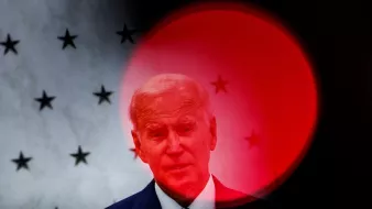 در این تصویر، شخصی در حال سخنرانی است و در پسزمینه، پرچم یا کاغذی با ستارههایی مشاهده میشود. به نظر میرسد که تصویر دچار افکت نوری شده و در مرکز، دایرهای قرمز رنگ وجود دارد که توجه را جلب میکند. این حالت میتواند حس خاصی به تصویر بدهد و بر حالت فرد تأکید کند.