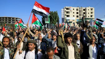 در این تصویر، تعداد زیادی از مردم در خیابانی تجمع کردهاند و پرچمهایی را در دست دارند. هر فردی ظاهری متعهد و با انرژی به نظر میرسد و برخی از آنها در حال فریاد زدن یا شعار دادن هستند. لباسها معمولاً سنتی هستند و برخی از شرکتکنندگان در حال بیان نظرات یا احساسات خود در مورد موضوعی خاص به نظر میرسند. محیط اطراف شامل ساختمانهای بلند و تابلوهای تبلیغاتی است که نشاندهنده یک میدان عمومی یا خیابان پرجنبوجوش است. پرچمها به رنگهای مختلف، خصوصاً رنگهای قرمز، سیاه و سفید، به خوبی دیده میشوند.