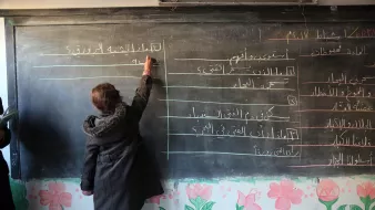 در این تصویر، یک دانشآموز در حال نوشتن روی تخته سیاه در کلاس درس است. او با دقت در حال نوشتن مطالبی است که به نظر میرسد مربوط به درس خاصی باشد. در پسزمینه، دیوار کلاس با نقاشیهای رنگی و گلها تزئین شده است. این فضا نمایانگر یک محیط آموزشی است که در آن دانشآموزان مشغول یادگیری هستند.