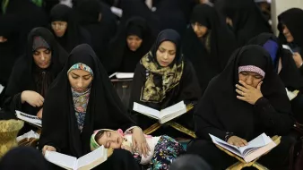 در تصویر گروهی از زنان با چادرهای سیاه در حال مطالعه قرآن دیده میشوند. بعضی از آنها در حال خواندن هستند و توجه عمیقی به کتاب دارند. همچنین، یک کودک در آغوش یکی از زنان خوابیده است. بهطور کلی، فضایی معنوی و پر از تمرکز در این تصویر وجود دارد.