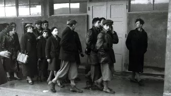 این تصویر سیاهوسفید گروهی از افراد را نشان میدهد که در یک محیط داخلی در حال حرکت هستند. برخی از افراد لباسهای گرم و کلوفت به تن دارند و به نظر میرسد که در حال عبور از یک فضای بسته هستند. تعدادی از افراد در حال گفتگو یا مراقب هستند، در حالی که دیگران به جلو نگاه میکنند. فضای تصویر حالت جدی و متفکری دارد و به طور کلی جو نگرانکنندهای را منتقل میکند.