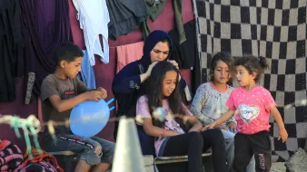 تصویر گروهی از کودکان و یک زن را نشان میدهد که در یک محیط غیررسمی نشستهاند. زن در حال بافتن یا شانه کردن موی دختری است که روی صندلی نشسته است. سایر کودکان دور آنها در حال بازی یا گفتگو هستند. پسزمینه تصویر به وضوح نشاندهنده محیطی ساده و روستایی است، با لباسهای آویخته به دیوار و برخی وسایل دیگر در اطراف. جو تصویر دوستانه و صمیمی به نظر میرسد.