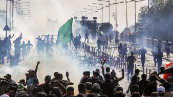 تصویر نشاندهنده یک تظاهرات یا اعتراض عمومی است که در یک خیابان یا پل در حال برگزاری است. تعداد زیادی از معترضان در حال حرکت و تجمع هستند و برخی از آنها پرچمهایی را در دست دارند. دود ناشی از گاز اشکآور یا مواد دیگر در هوا دیده میشود و با ماشینهای مخصوص پلیس در پسزمینه، فضایی تنشآلود به وجود آمده است. نوارهای موانعی نیز در اطراف معترضان مشاهده میشود که برای کنترل جمعیت و جلوگیری از پیشروی آنها استفاده میشود.