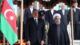 این تصویر دیداری رسمی را نشان میدهد که در آن دو فرد به همراه یکدیگر ایستادهاند. یکی از آنها در لباس رسمی و دیگری در لباس روحانی به چشم میخورد. در پسزمینه پرچم آذربایجان دیده میشود و تعدادی از افراد دیگر نیز در پسزمینه حاضرند. فضای کلی تصویری رسمی و دولتی به نظر میرسد.