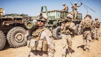 در این تصویر، گروهی از سربازان نظامی در حال فعالیت در یک محیط بیابانی دیده میشوند. آنها لباسهای نظامی به تن دارند و بخشی از تجهیزات سنگین را بارگیری میکنند. یکی از سربازان در پیشزمینه، موشک یا سلاحی را به دوش گرفته و به سمت دوربین نگاه میکند. در پسزمینه، وسیله نقلیه نظامی و دیگر سربازان مشغول به کار در حال حرکت هستند. آسمان آبی و صاف به همراه زمین خشک و شنزار نمایی از یک منطقه نظامی را به تصویر میکشد.