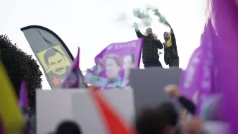 در این تصویر، تعدادی از افراد در حال برگزاری یک تجمع اعتراضی یا حمایتی به نظر میرسند. دو نفر بر روی یک سکو ایستادهاند و یکی از آنها میکروفون در دست دارد و به جمعیت صحبت میکند. در دست برخی از افراد پرچمها و بنرهایی با رنگهای مختلف، از جمله بنرهای بنفش، در حال تکان خوردن است. همچنین، دود سبز رنگی به هوا بلند شده که به جو تجمع حال و هوای خاصی میبخشد. در پسزمینه، تابلوی چهرهای نیز قابل مشاهده است.