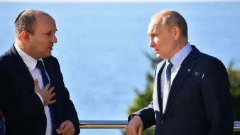 در این تصویر، دو مرد در حال گفتگو هستند. یکی از آنها کت و شلوار تیره به تن دارد و به سمت دیگر شخص اشاره میکند. مرد دیگر نیز کت و شلوار رسمی پوشیده و به سمت شخص اول نگاه میکند. پسزمینه تصویر نیز شامل دریا و آسمان است که فضایی آرامشبخش را ایجاد کرده است.