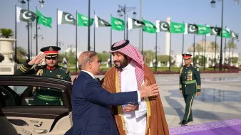 دو مرد در لباسهای رسمی در یک محیط بیرونی با پرچمهای پاکستان در پسزمینه به هم خوشآمد میگویند.