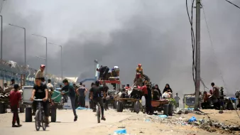 تصویر یک صحنه شهری را نشان میدهد که در آن تعدادی از افراد در حال حرکت هستند. در زمینه، دود سیاهی به آسمان بلند شده که نشاندهنده وجود یک حادثه یا آتشسوزی است. افرادی که در تصویر دیده میشوند، شامل مردان، زنان و کودکانی هستند که برخی از آنها در حال دوچرخهسواری و برخی دیگر در حال حمل بار در کالسکههای دستی هستند. فضای کلی تصویر به نظر شلوغ و پر از فعالیت است.