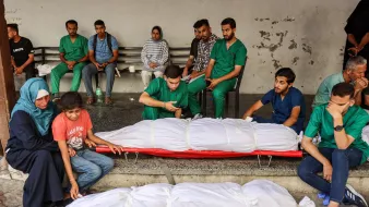 در این تصویر، تعدادی از افراد در یک مکان عمومی نشستهاند. در جلو، دو بدن پوشیده شده در پارچههای سفیدی قرار دارند. افراد حاضر در تصویر با ظاهری نگران و غمگین به نظر میرسند. برخی از آنها لباسهای پزشکی به تن دارند و به وضوح تحت تأثیر شرایط عاطفی و وضعیت حاضر قرار گرفتهاند. فضا به طور کلی حاکی از یک وضعیت بحرانی و دردناک است.