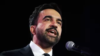 مردی با لباس رسمی، در حال سخنرانی و صحبت در میکروفون است. احساس جدیت و اعتماد به نفس دارد.